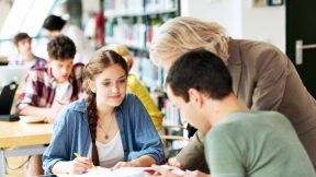 Internationale Studierende sitzen an Tischen in der Bibliothek, eine Dozentin hilft beim Lernen.